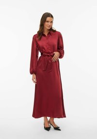 Bordowa sukienka maxi z satyny z kołnierzykiem, długimi bufkami na rękawach, zapięciem na guziki z przodu i przewiązanym w talii paskiem. Charakteryzuje się gładką fakturą i swobodnym krojem.