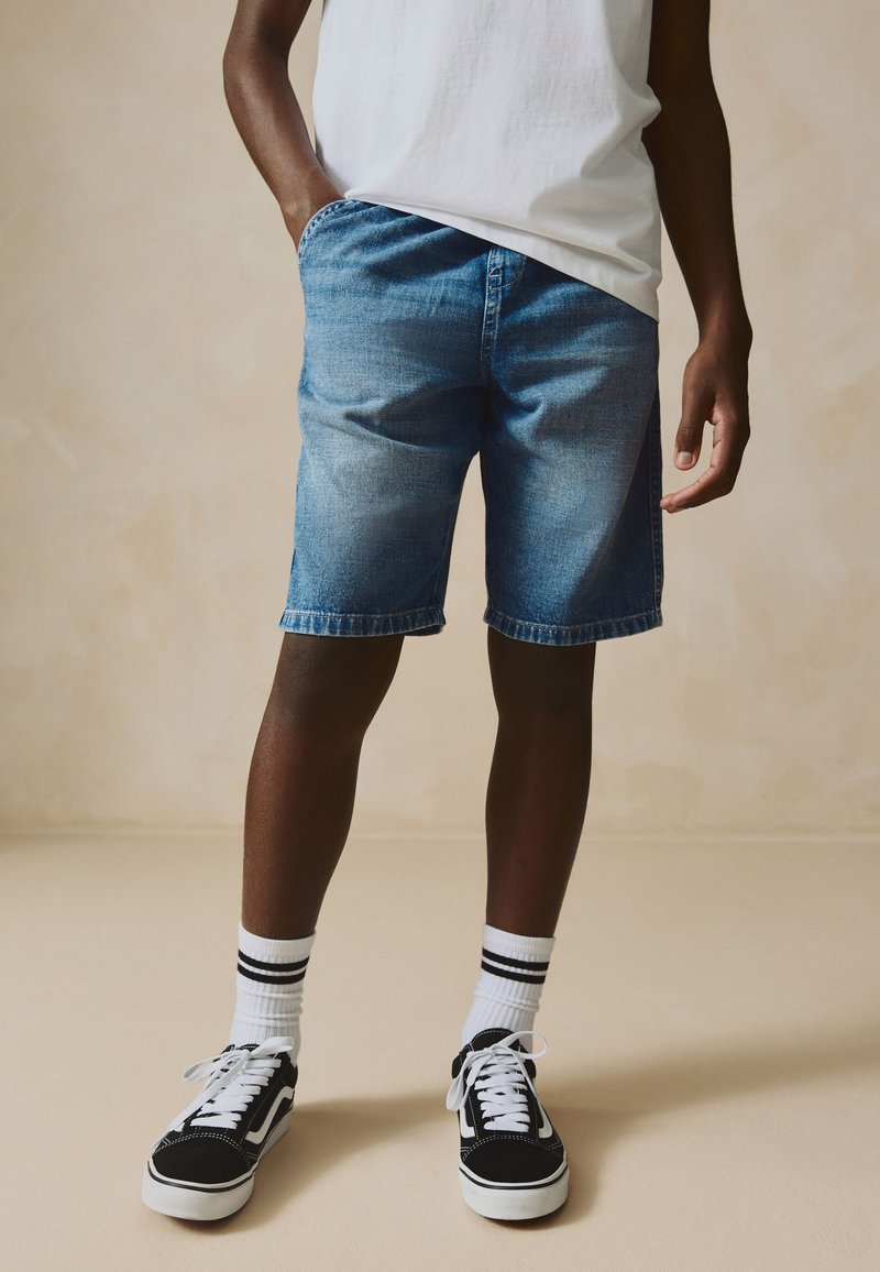 Mand iført lyseblå denimshorts, hvid T-shirt, hvide sokker med sorte striber og sorte og hvide sneakers, stående mod en beige baggrund.