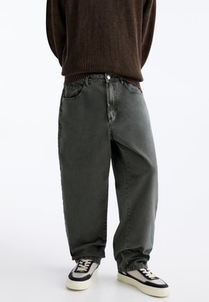 Person trägt dunkelgraue, weit geschnittene Jeans, braunen gerippten Pullover und schwarz-weiße Sneaker, steht vor einem weißen Hintergrund.