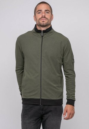 Homme portant une veste zippée vert olive avec col et poignets noirs, debout devant un fond gris clair uni.