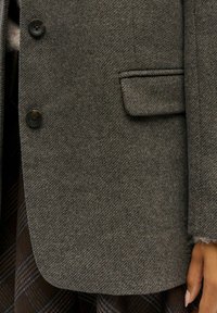 Grau-gestreifter Woll-Blazer mit zwei schwarzen Knöpfen, einer einzelnen Brusttasche und einer weichen Textur. Über einem karierten Rock getragen.