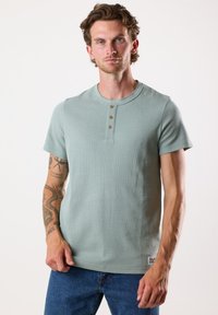 Ljusgrön kortärmad henley-t-shirt med tre bruna knappar vid halsringningen, tillverkad av texturerat tyg; kombinerad med blå jeans i denim.