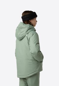 Πράσινο επενδεδυμένο μπουφάν με κουκούλα, υφασμάτινη υφή, πλαϊνές τσέπες, και μαύρη πλεκτή κορδέλα κεφαλής που φοράει ένα πρόσωπο που είναι γυρισμένο με την πλάτη.