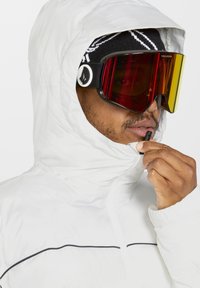 Witte geïsoleerde jas met zwarte accenten, voorzien van een capuchon en ritssluiting. Reflecterende skibril met een gradient lens in rood en geel.