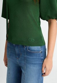Top verde dalla texture con maniche corte a palloncino, caratterizzato da un piccolo logo dettagliato all'orlo, abbinato a jeans in denim blu.