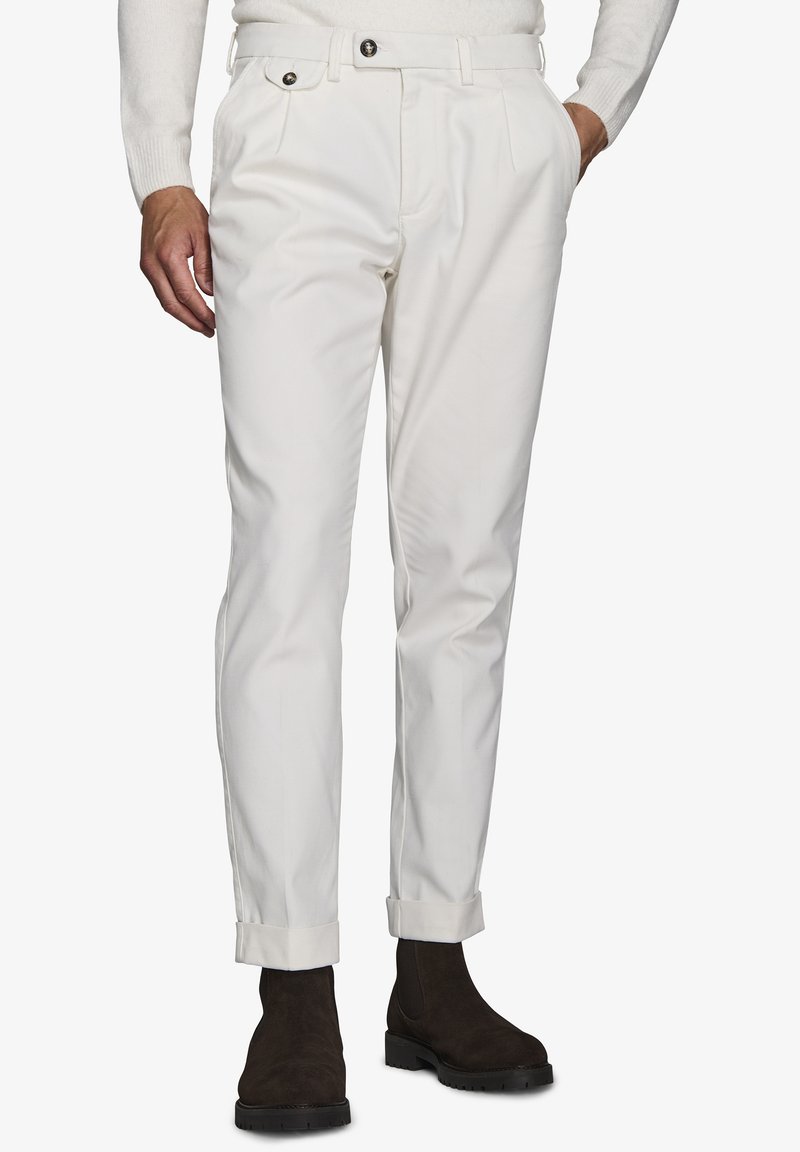 Weiße Baumwollhose mit Slim-Fit, gefalteter Vorderseite und umgeschlagenen Säumen, kombiniert mit braunen Wildlederstiefeln. Zwei schwarze Knöpfe am Bund.