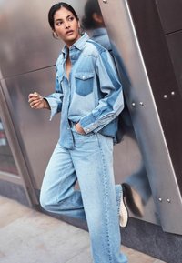 Camicia di denim in tonalità azzurro chiaro, con un taglio oversize, taschino sul petto e chiusura con bottoni. Abbinata a jeans azzurro chiaro a vestibilità ampia e scarpe bianche.