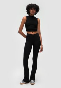 Černý crop top s úzkým střihem a vysokým výstřihem, ve spojení s černými rozšířenými legínami. Model má na sobě metalické stříbrné boty.