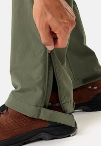 Grüne Outdoor-Hosen mit seitlichen Reißverschlüssen, kombiniert mit braunen Wanderschuhen mit strukturierten Sohlen und schwarzen Akzenten.