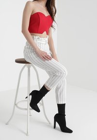 Kvinna som sitter på en vit metallpall, iklädd en röd ärmlös topp, vita randiga högmidjade byxor och svarta stövletter med klack.