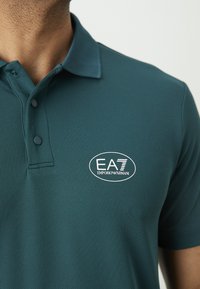 Vyriškis dėvi smaragdo spalvos marškinėlius su apykakle ir dviem mygtukais, o krūtinėje matomas baltas EA7 Emporio Armani logotipas.