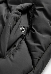 Schwarze Pufferjacke aus Stoff mit gestepptem Muster, glänzenden schwarzen Druckknöpfen und verstärkten Nähdetails. Aus weichem, isoliertem Material.
