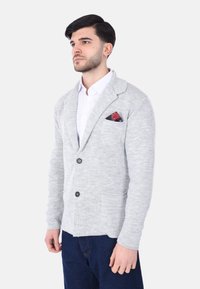 Cardigan grigio a maglia con colletto a scialle, due tasche frontali e bottoni neri. Presenta un fazzoletto rosso a motivo nella tasca sul petto sinistro.