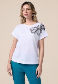 Witte t-shirt met korte mouwen, versierd met een glinsterende zwarte strikaccent op de linker schouder; gecombineerd met teal gekleurde broeken. Gemaakt van zacht katoen.