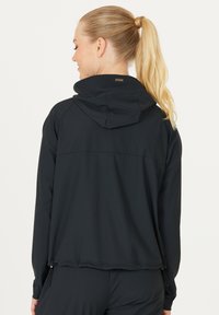 Giacca sportiva nera con cappuccio, polsini a costine e orlo arricciato. Presenta un tessuto leggero e liscio e un piccolo dettaglio del logo sul retro.