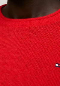 Roter Strickpullover mit geripptem Rundhalsausschnitt, strukturierter Oberfläche und einem kleinen Logo-Akzent in Blau, Weiß und Rot an der Seite.
