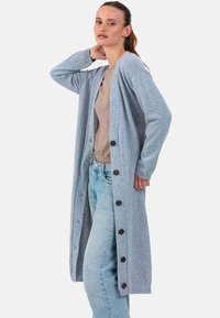 Cardigan long bleu clair avec poignets côtelés et boutons en bois, assorti à un top beige et un jean bleu clair. Tissu doux et texturé.