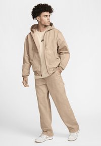 Brun tät hoodie med framfickor, lager på en ljus beige t-shirt med en liten svart logotyp; parat med matchande lösa byxor och vita sneakers.