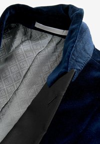Col de veste en velours bleu marine avec revers noirs et doublure en soie gris clair à motifs, montrant les textures détaillées du tissu et les coutures.