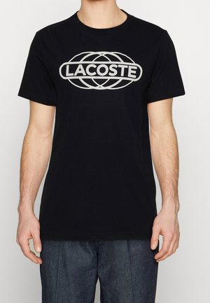 T-shirt en coton noir affichant un grand logo Lacoste blanc en design géométrique sur la poitrine. Col rond et manches courtes.