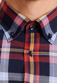 Zbliżenie na szyję osoby ubraną w koszulę w kratkę w kolorze czerwonym, granatowym i żółtym z kołnierzykiem zapinanym na guziki.