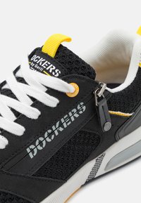 Baskets noires avec lacets blancs, en maille et matériaux synthétiques, avec une languette jaune, des éléments en métal argenté, et le logo "DOCKERS" imprimé sur le côté.