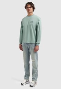 Jonge man die staat te gekleed in een mintgroene langemouwen shirt, lichte verwasemde jeans en zilveren hardloopschoenen op een effen lichte achtergrond.