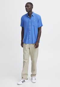 Camisa azul de manga corta con bolsillo, combinada con pantalones beige claros y zapatillas blancas con rayas negras. Diseño simple y casual.
