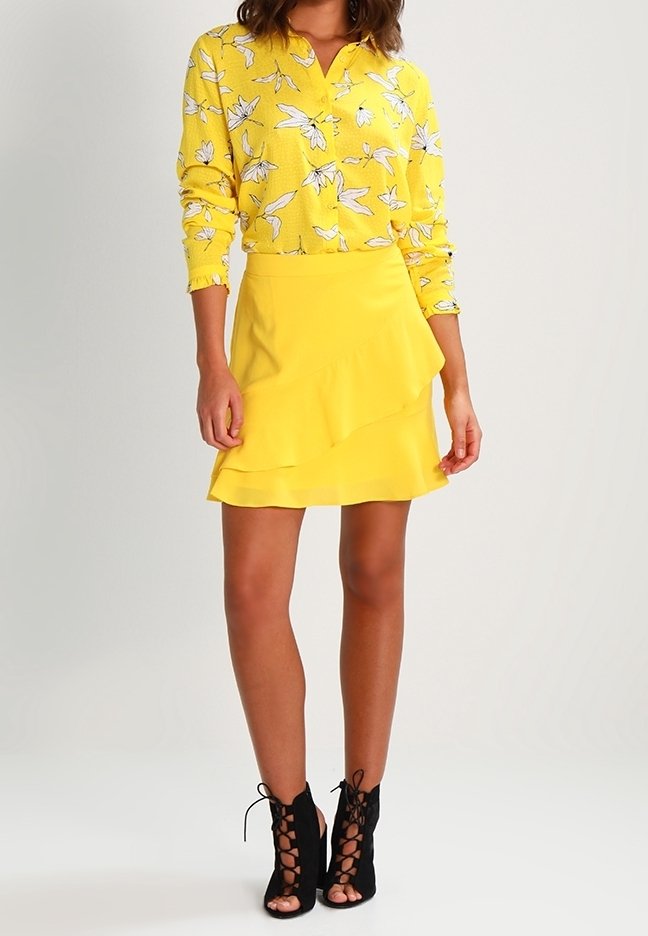 Gelbe Bluse mit weißem Blumenmuster, kombiniert mit einem gestuften gelben Rock. Schwarze Schnürstiefeletten runden das Outfit ab.