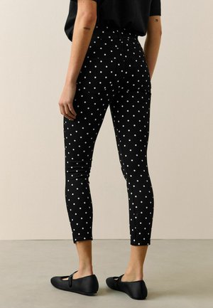 Person trägt schwarze, eng anliegende Hosen mit weißen Punkten und schwarze Mary-Jane-Ballerinas, steht vor einem neutralen Hintergrund und blickt weg.