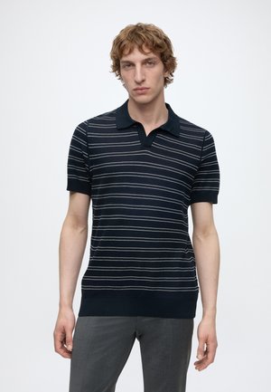 Tânăr cu păr ondulat castaniu deschis, purtând un tricou polo albastru închis cu mâneci scurte, cu dungi albe orizontale subțiri și pantaloni gri.