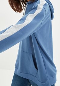 Persona vestida con sudadera con capucha azul y franjas blancas en las mangas y el bolsillo delantero, combinada con jeans azules, contra un fondo liso.