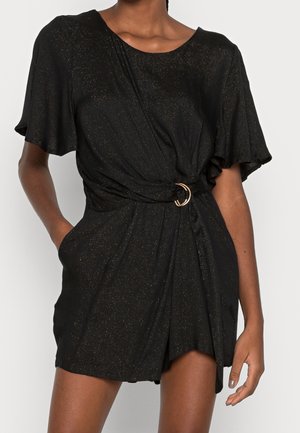 Femme portant une combinaison-short noire pailletée à manches courtes, avec un devant drapé et une ceinture à boucle dorée à la taille.