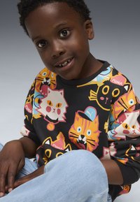 Sudadera negra con patrones de gatos coloridos, que presenta varias caras en estilo de caricatura. Usada con jeans de color azul claro, en una pose sentada.