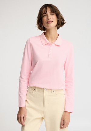 Frau mit kurzen braunen Haaren, die ein hellrosa langärmeliges Poloshirt und beige Hose vor weißem Hintergrund trägt.