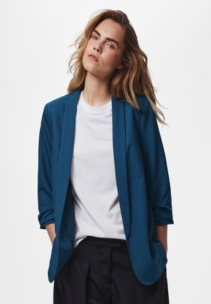 Ung person med langt hår iført en blå blazer over en hvid t-shirt og sorte bukser, står med hænderne i lommerne mod en ensfarvet baggrund.