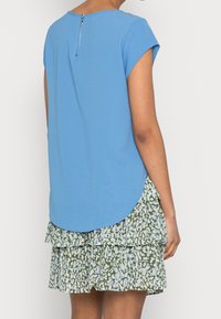 Top azul de manga corta con dobladillo redondeado y detalle de cremallera en la parte posterior, superpuesto sobre una falda de capas en tonalidades verdes y azules con un patrón.