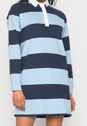 Mujer vestida con un vestido de rugby de manga larga con amplias franjas horizontales en azul marino y azul claro y un cuello blanco abotonado.