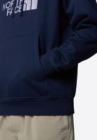 Sudadera azul marino con bolsillo frontal, dobladillo y puños acanalados, con un logo blanco en el pecho. Textura suave, ajuste casual.