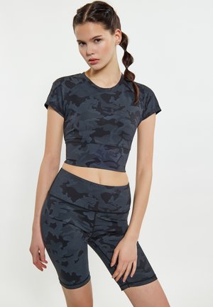 Jonge vrouw met gevlochten haar, gekleed in een bijpassende donkergrijze camo crop top en biker shorts, poserend tegen een witte achtergrond.
