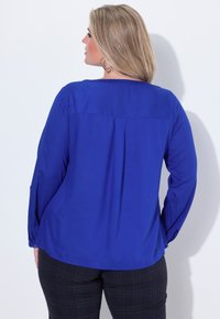 Blusa blu a maniche lunghe con retro plissettato, realizzata in tessuto morbido. Abbinata a pantaloni a quadri scuri con un motivo a griglia marrone.