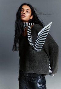 Pulover tricotat de culoare verde închis, cu o croială lejeră, purtat deasupra unei cămăși în dungi alb-negru. Pantaloni din piele negri vizibili la talie.