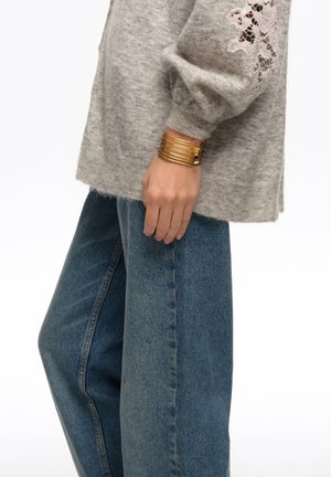 Person trägt blaue Jeans mit weitem Bein und einen grauen Pullover mit Spitzen-Details an den Ärmeln sowie mehrere goldene Armreifen am Handgelenk.