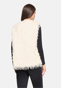 Kremowa kamizelka z futerka sztucznego o długiej, puszystej fakturze, z wysokim kołnierzem i otwartym przodem. Noszona na czarnej bluzce z długim rękawem.