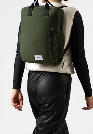 Osoba ubrana w czarne długie rękawy, kremową kamizelkę i czarne spodnie niesie kwadratowy zielony plecak z przednią kieszenią na suwak i czarnymi uchwytami.