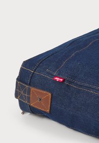 Tumši zila denima soma ar brūnu ādas akcentu, ar dzeltenām diega pērlēm, dekoratīvo uzlīmi un mazu sarkanu zīmola etiķeti.