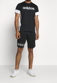 Musta adidas T-paita, jossa valkoinen logo ja sivuakkentsit, yhdistettynä mustiin shortsihousuihin, joissa on "adidas"-teksti, ja valkoisiin urheilukenkiin.