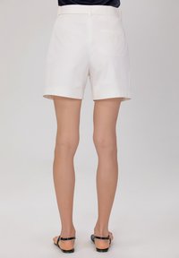 Shorts en coton blanc avec une taille plate, des poches latérales et arrière, des ourlets retroussés et une texture lisse, associés à des sandales noires.
