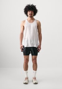 White, sleeveless, perforated tank top with a side pocket, paired with black shorts featuring red accents. Model stands against a plain background.