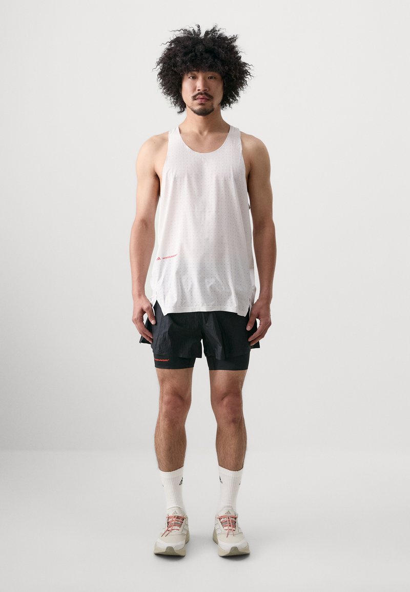 White, sleeveless, perforated tank top with a side pocket, paired with black shorts featuring red accents. Model stands against a plain background.